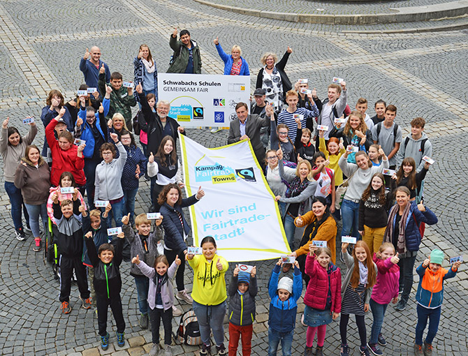 Alle Beteiligten halten eine Fairtrade-Fahne hoch und freuen sich.