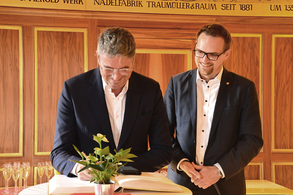 Zwei Männer in dunklen Anzügen und weißem Hemd stehen im Goldenen Saal und geben sich die Hand. Vor ihnen liegt aufgeschlagen das Goldene Buch.