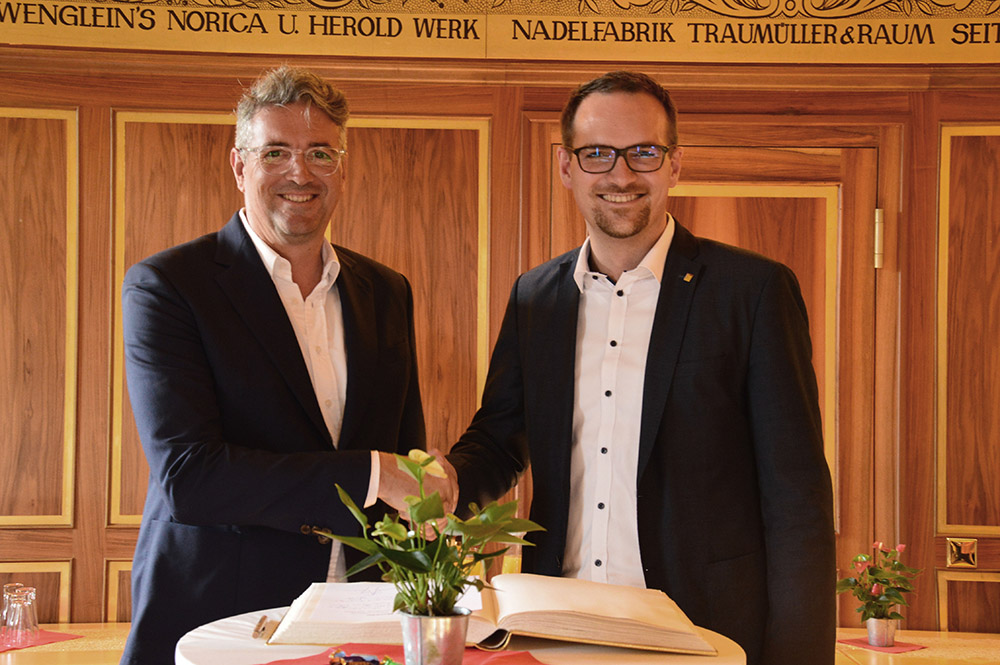 Zwei Männer in dunklen Anzügen und weißem Hemd stehen im Goldenen Saal und geben sich die Hand. Vor ihnen liegt aufgeschlagen das Goldene Buch.