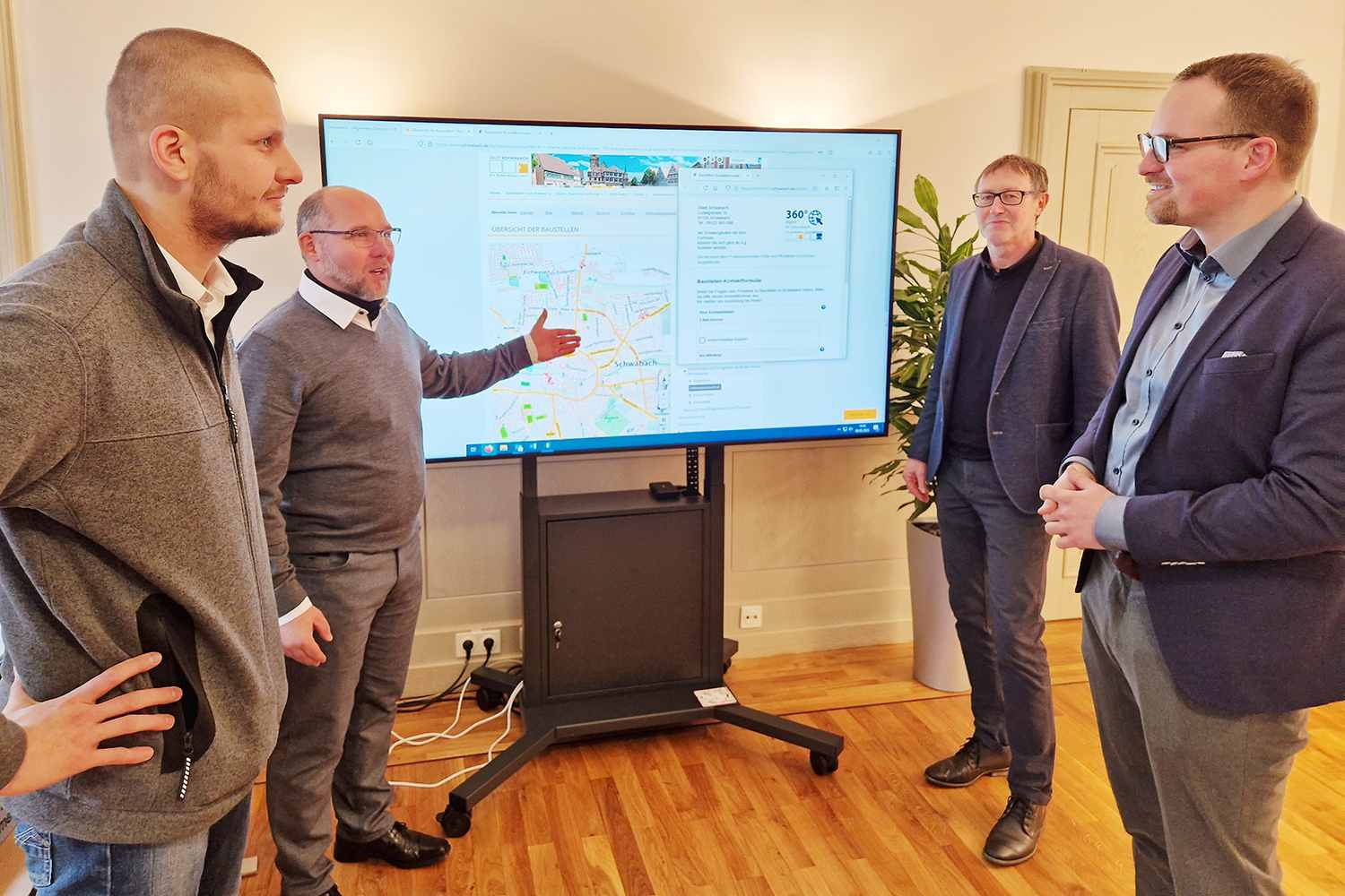 Vier Männer stehen vor einem großen Bildschirm, darauf zu sehen ist eine Karte von Schwabach, die Baustellen sind durch rote Symbole markiert.