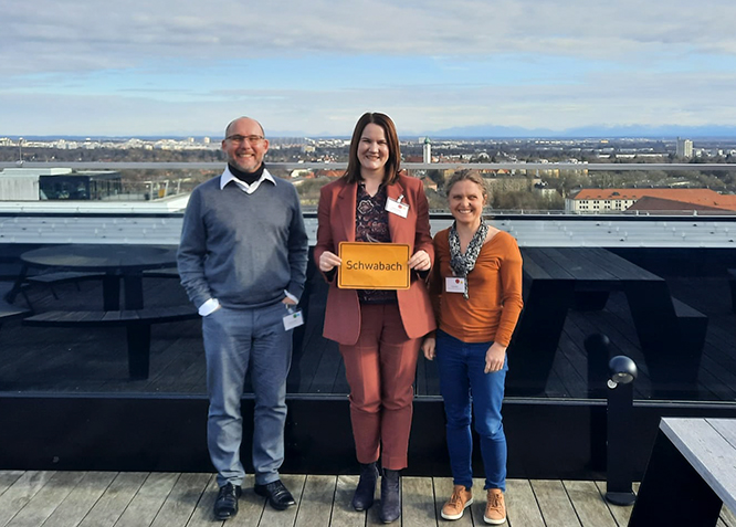 Ein Mann und zwei Frauen stehen auf einer Dachterrasse, die Frau in der Mitte hält ein gelbes Ortsschild "Schwabach" im A4-Format, dahinter ist die Skyline Münchens zu sehen.