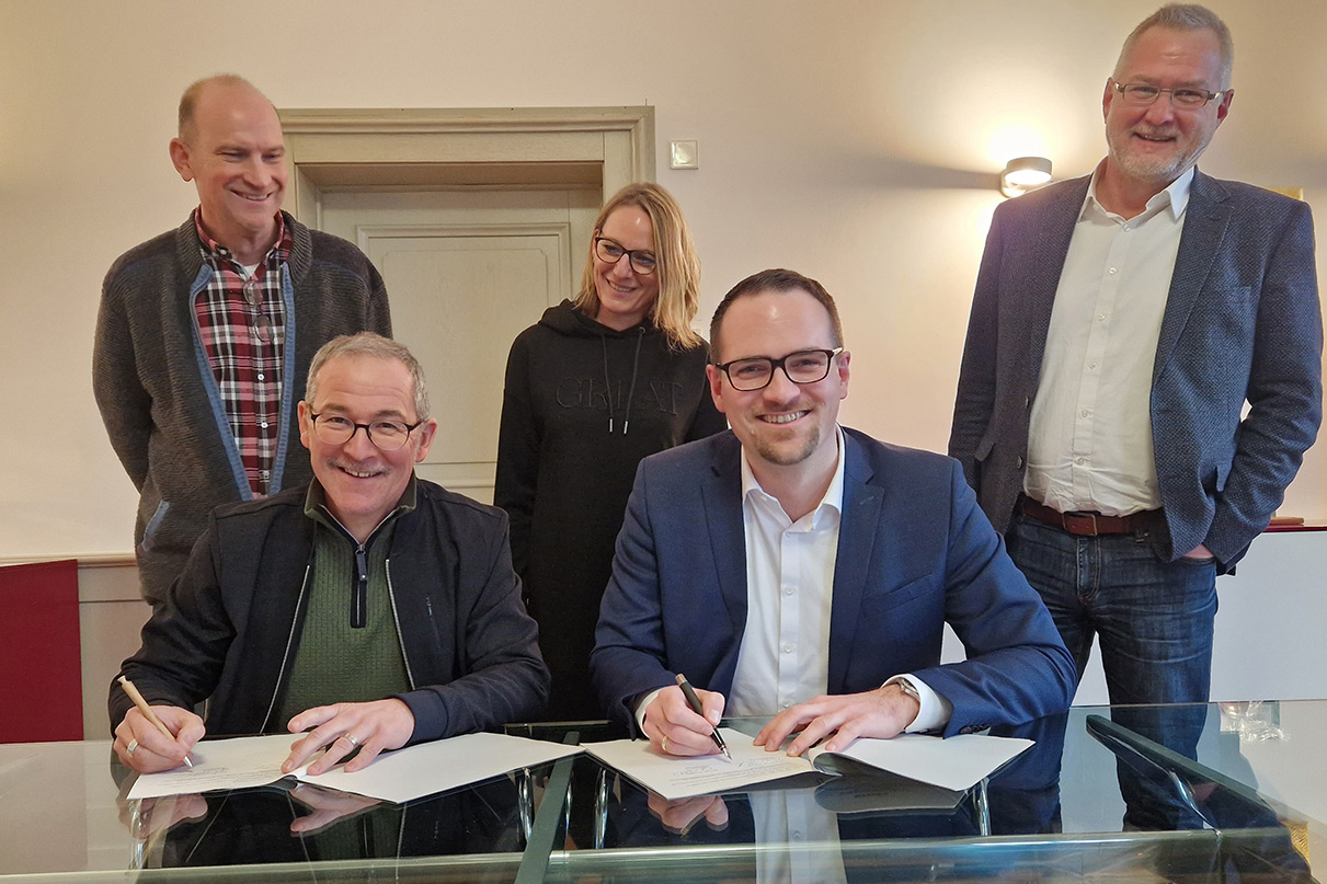 An einem Glastisch sitzen zwei Männer, die beiden Bürgermeister, und unterzeichnen die Zweckvereinbarung. Dahinter stehen zwei Männer und eine Frau, die zuständigen Mitarbeiter der beiden Verwaltungen.