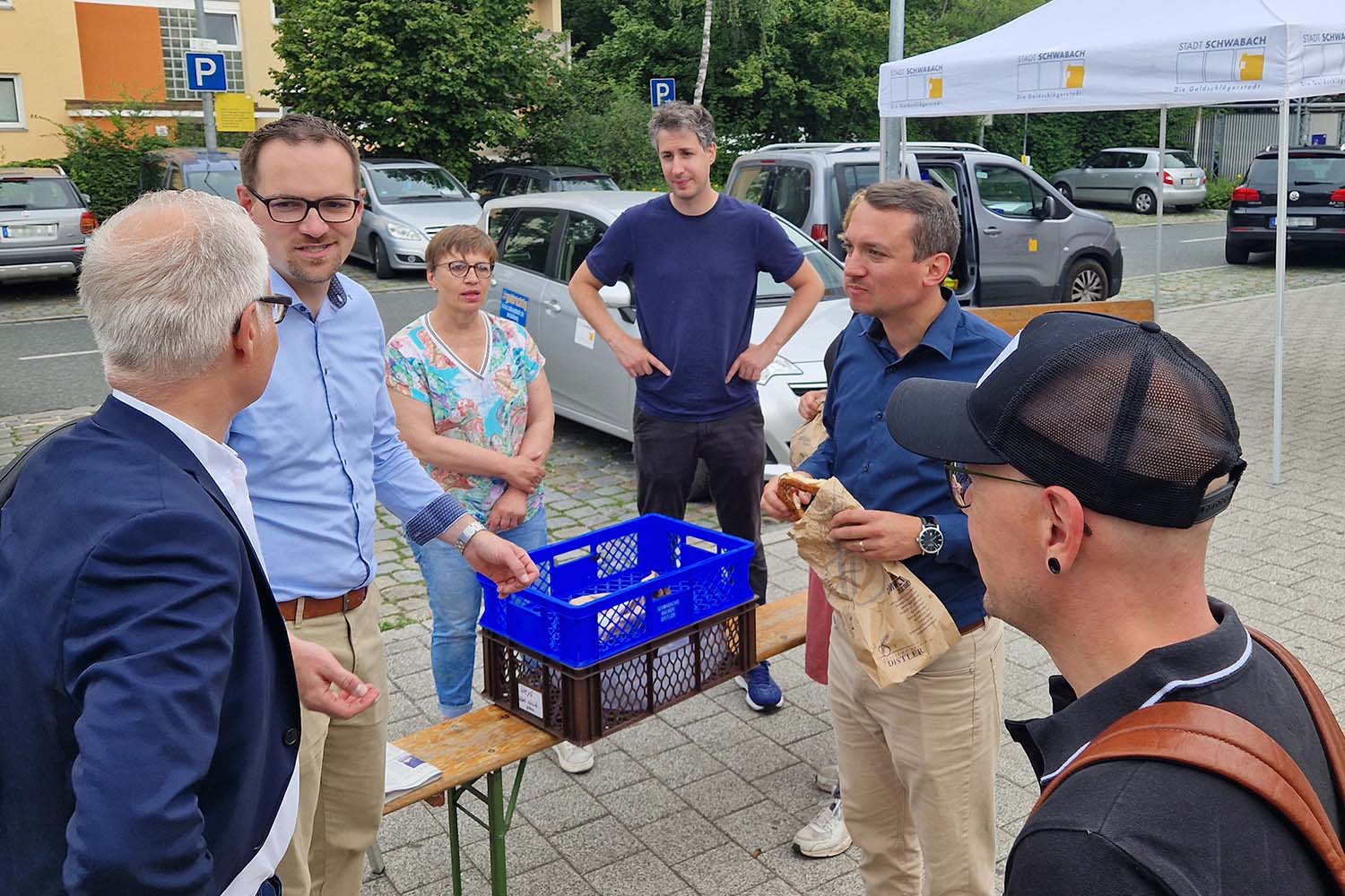 Mehrere Personen stehen um einen blauen Kasten, in dem Frühstückstüten liegen. Ein Mann hält eine Bäckertüte und eine Breze in der Hand.